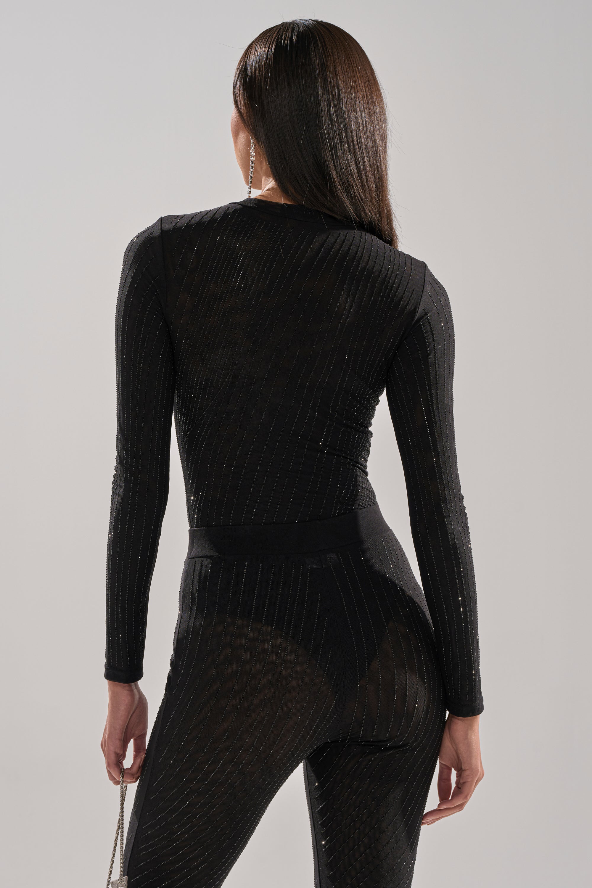 A woman with long brown hair faces away, holding a small bag, and wears the TWINKLING LIGHTS Rhinestone Long Sleeve Mesh Bodysuit in Black featuring vertical stripes. The background is plain and light-colored.