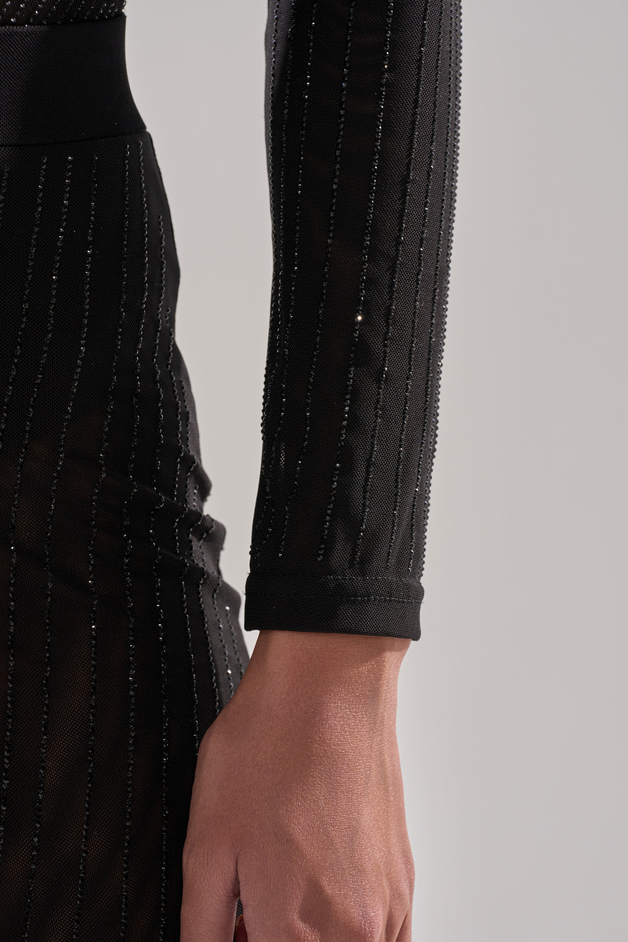 A close-up of a person's arm and hand in the TWINKLING LIGHTS RHINESTONE LONG SLEEVE MESH BODYSUIT IN BLACK, featuring shimmering vertical stripes. The background is plain and light-colored.