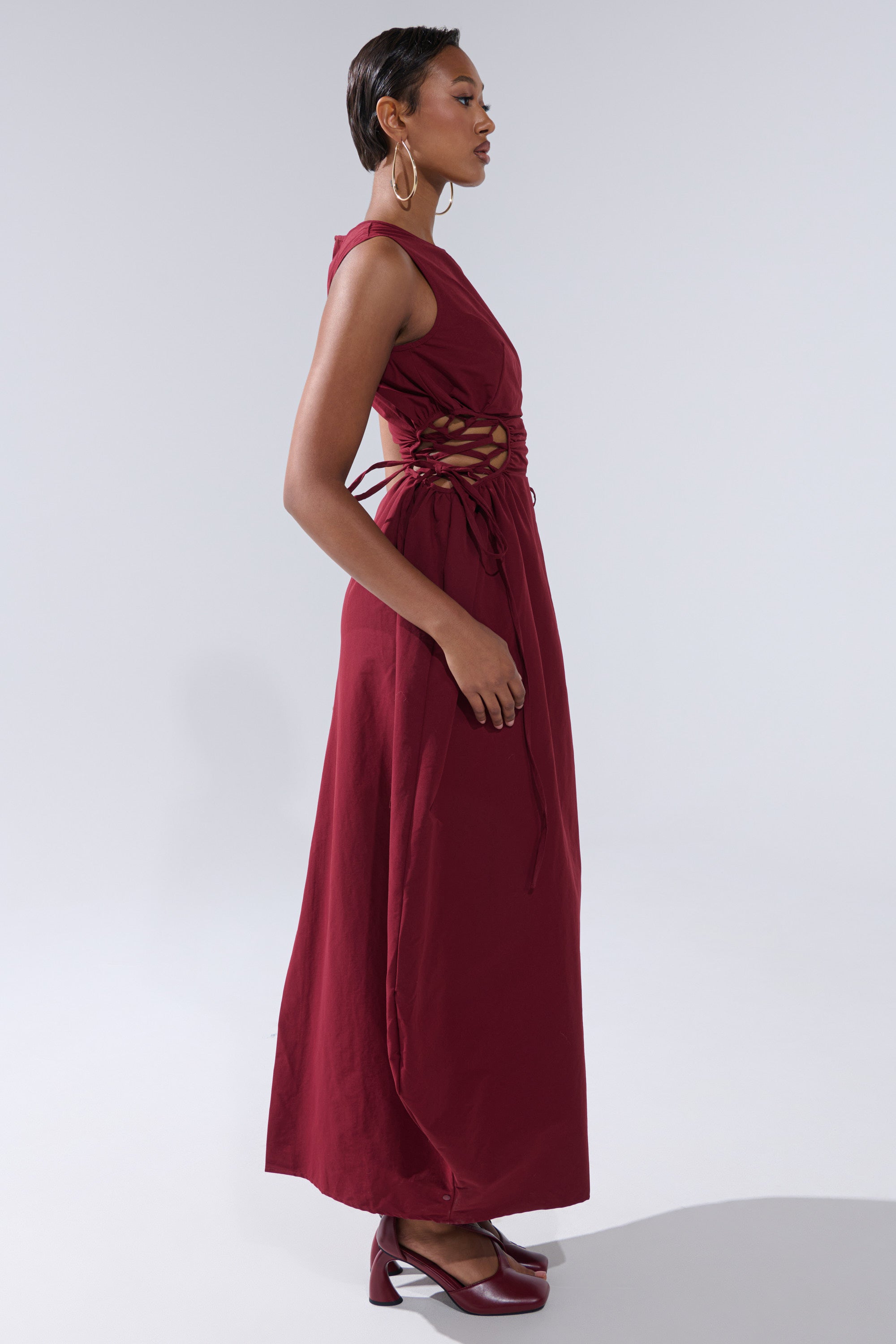 A woman in profile wears the ALINA LACE UP DETAIL MAXI DRESS IN BURGUNDY, a sleeveless, ankle-length dress with lace-up sides. She pairs it with matching shoes, has short slicked-back hair and large hoop earrings, set against a plain gray background.