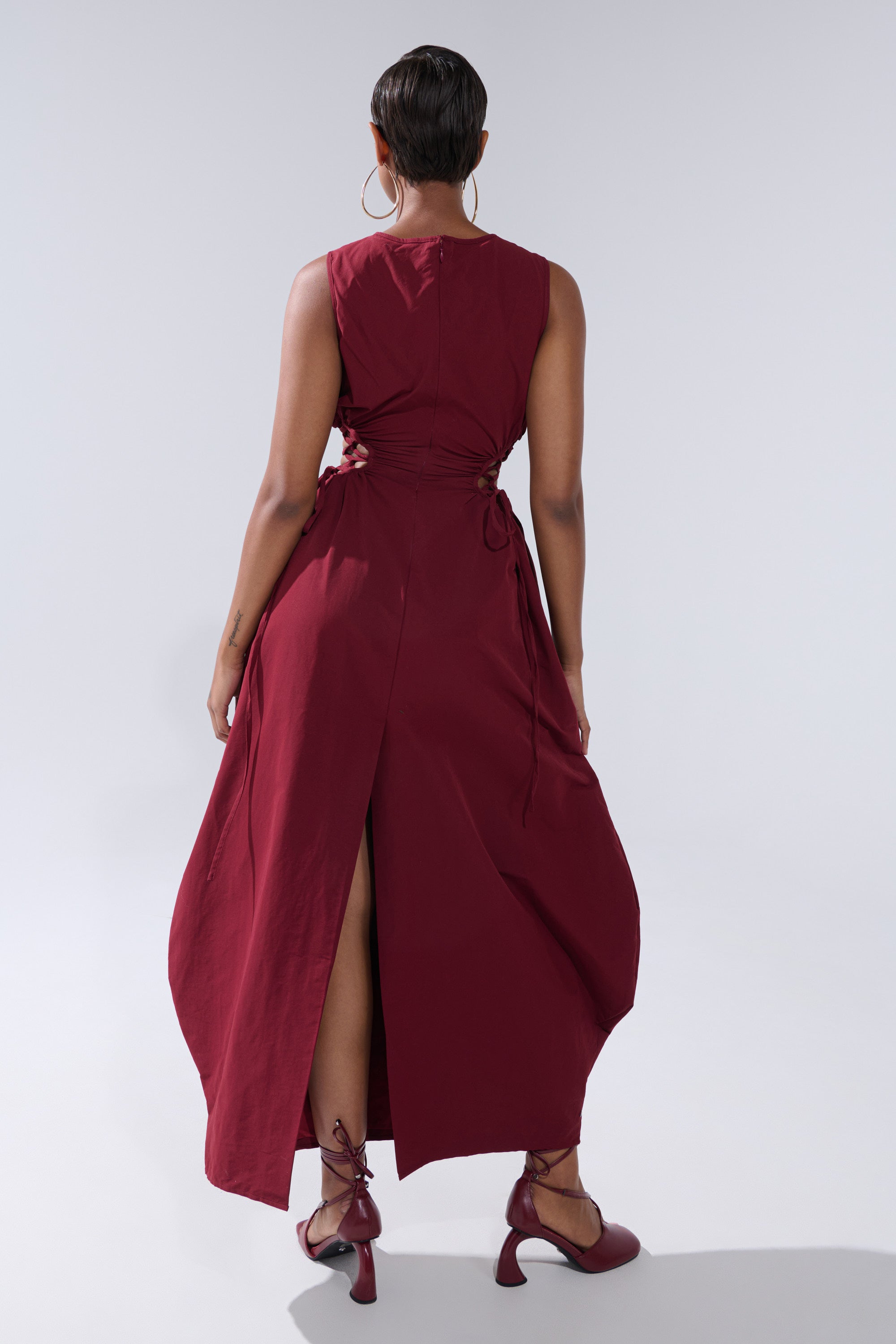 A person with short hair and large hoop earrings stands facing away, showcasing the ALINA LACE UP DETAIL MAXI DRESS IN BURGUNDY’s flowy fabric, open back, side ties, and an elegant back slit.