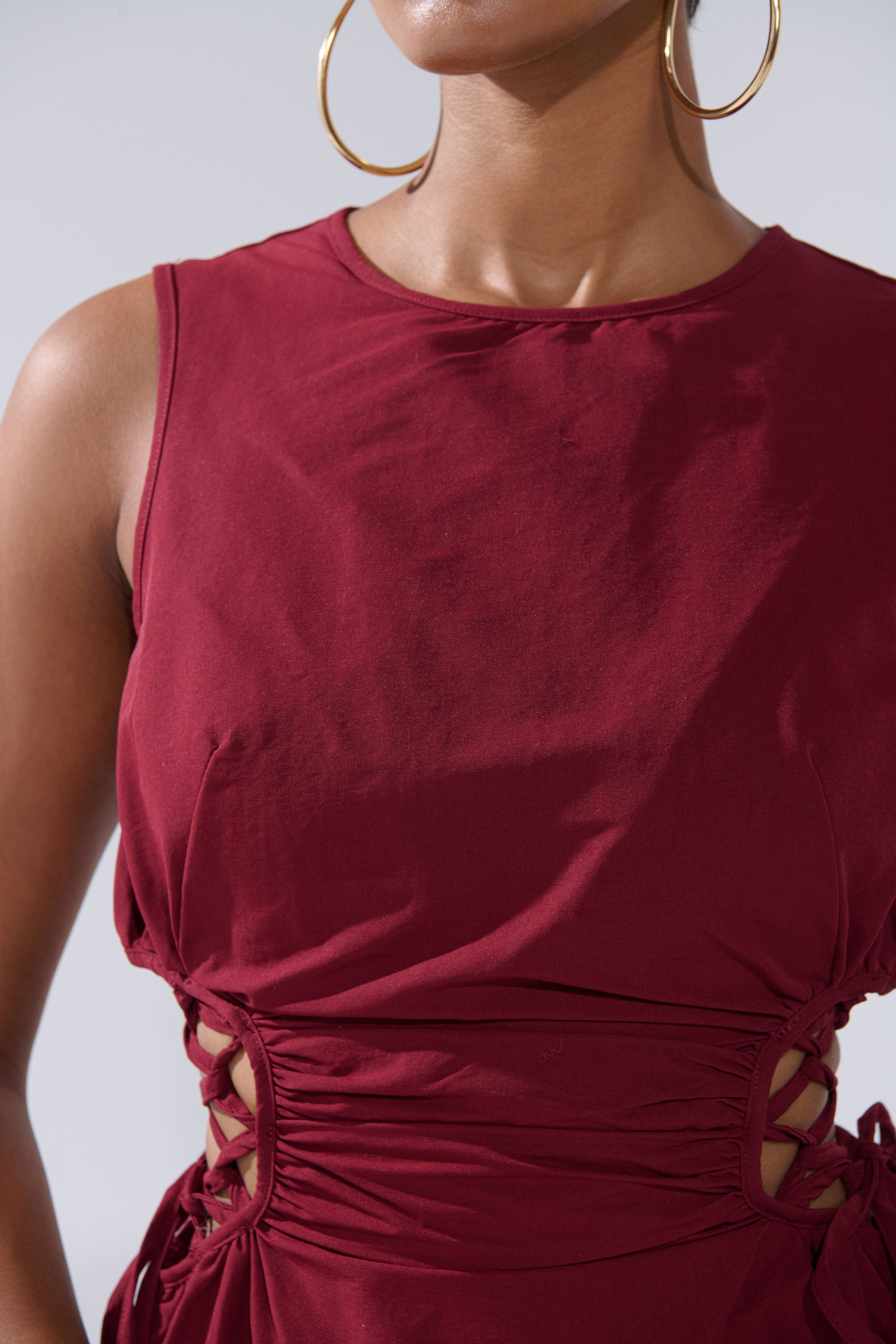 A close-up of a person wearing the ALINA LACE UP DETAIL MAXI DRESS IN BURGUNDY, a sleeveless maxi dress with side lace-up details, styled with large gold hoop earrings. The person's face is partially out of frame.