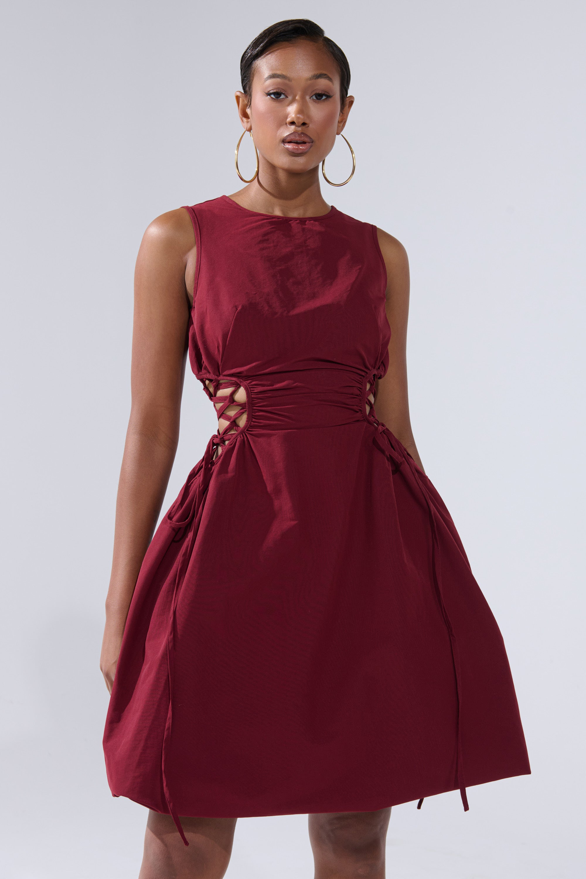 A woman with short hair and large hoop earrings stands confidently against a plain background, wearing the ALINA LACE UP DETAIL MAXI DRESS IN BURGUNDY, a sleeveless maxi dress featuring stylish side lace-up details.