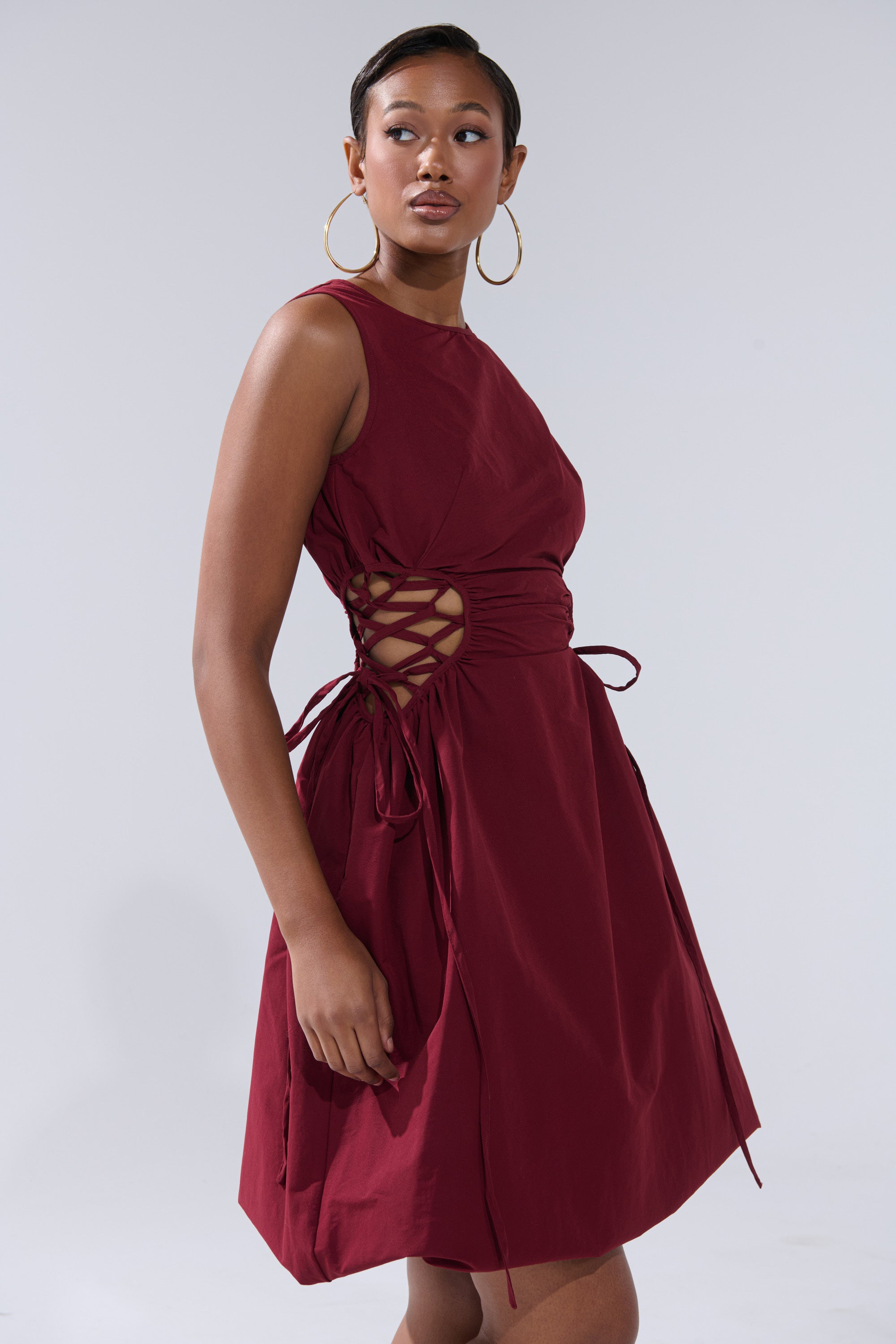 A woman in the ALINA LACE UP DETAIL MAXI DRESS IN BURGUNDY with lace-up sides and large hoop earrings stands against a light gray background, glancing over her shoulder.