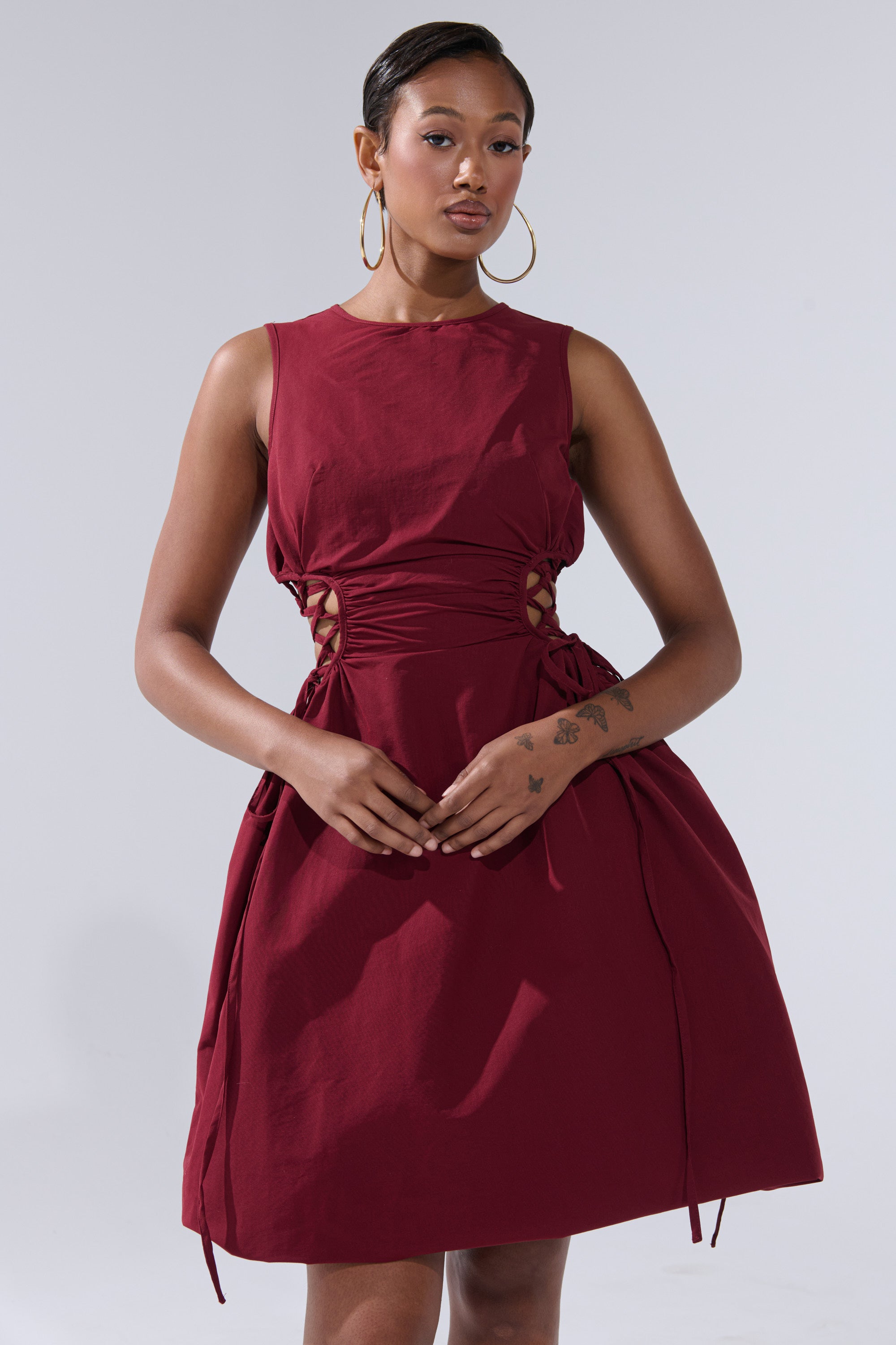 A woman with short hair, large hoop earrings, and visible left forearm tattoos poses against a light gray background wearing the sleeveless ALINA LACE UP DETAIL MAXI DRESS IN BURGUNDY, featuring side cutouts and ruched detailing.