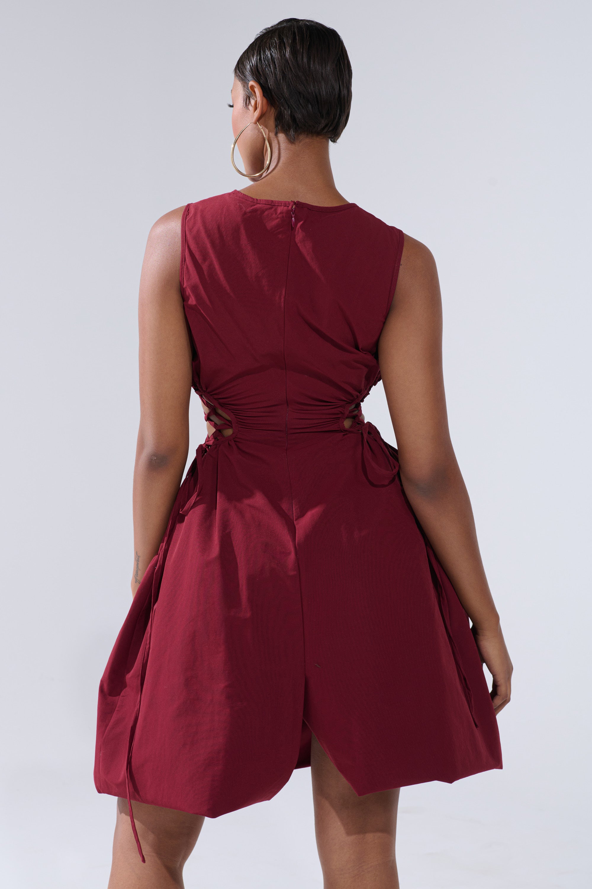 A person with short dark hair is shown from behind, wearing the ALINA LACE UP DETAIL MAXI DRESS IN BURGUNDY. The sleeveless dress features waist cutouts, a flared skirt, and is styled with large hoop earrings.
