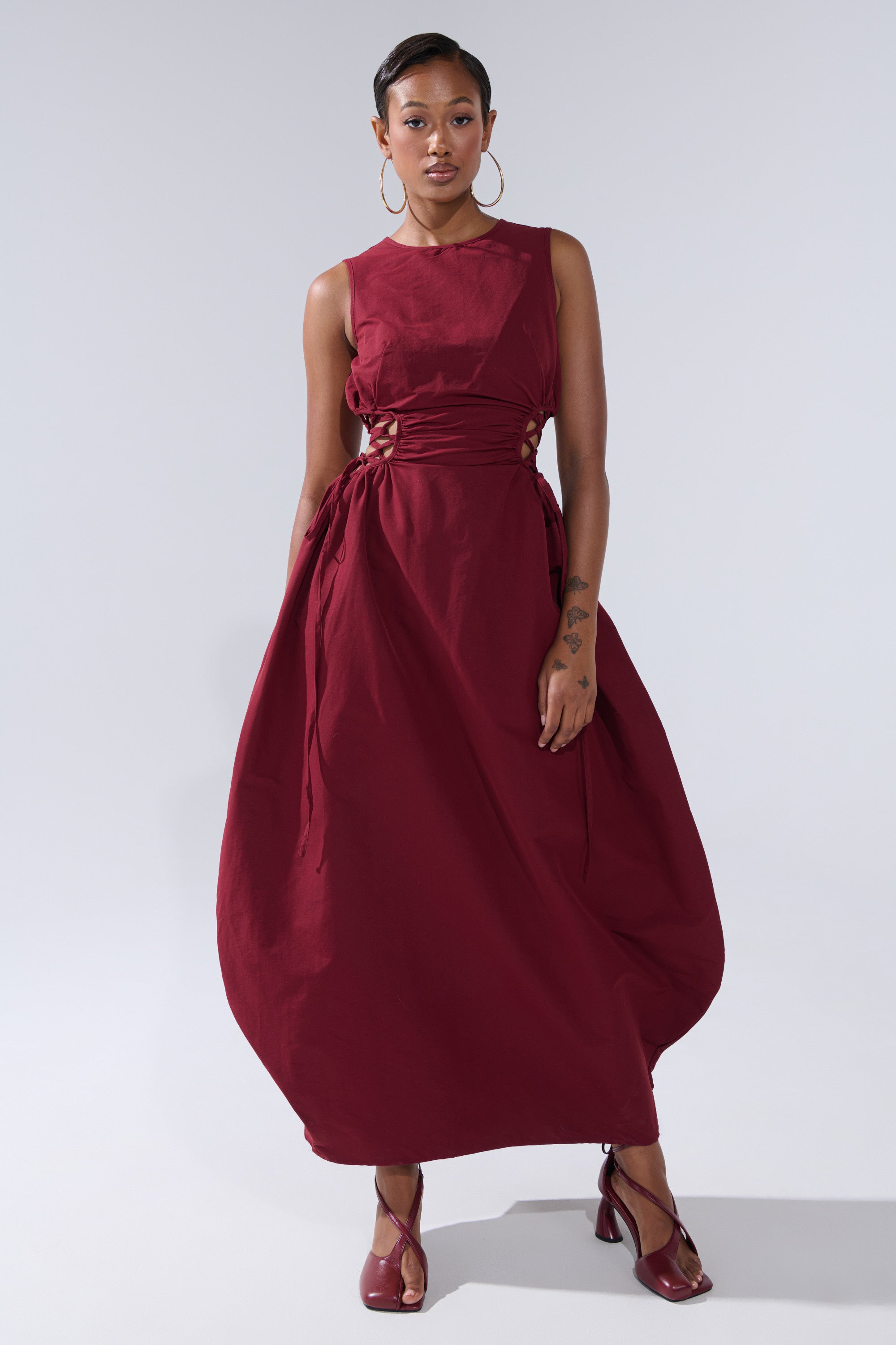 A woman stands confidently in the ALINA LACE UP DETAIL MAXI DRESS IN BURGUNDY, featuring sleeveless styling and side cutouts with ties. She completes her look with hoop earrings, maroon heels, and a forearm tattoo against a plain background.
