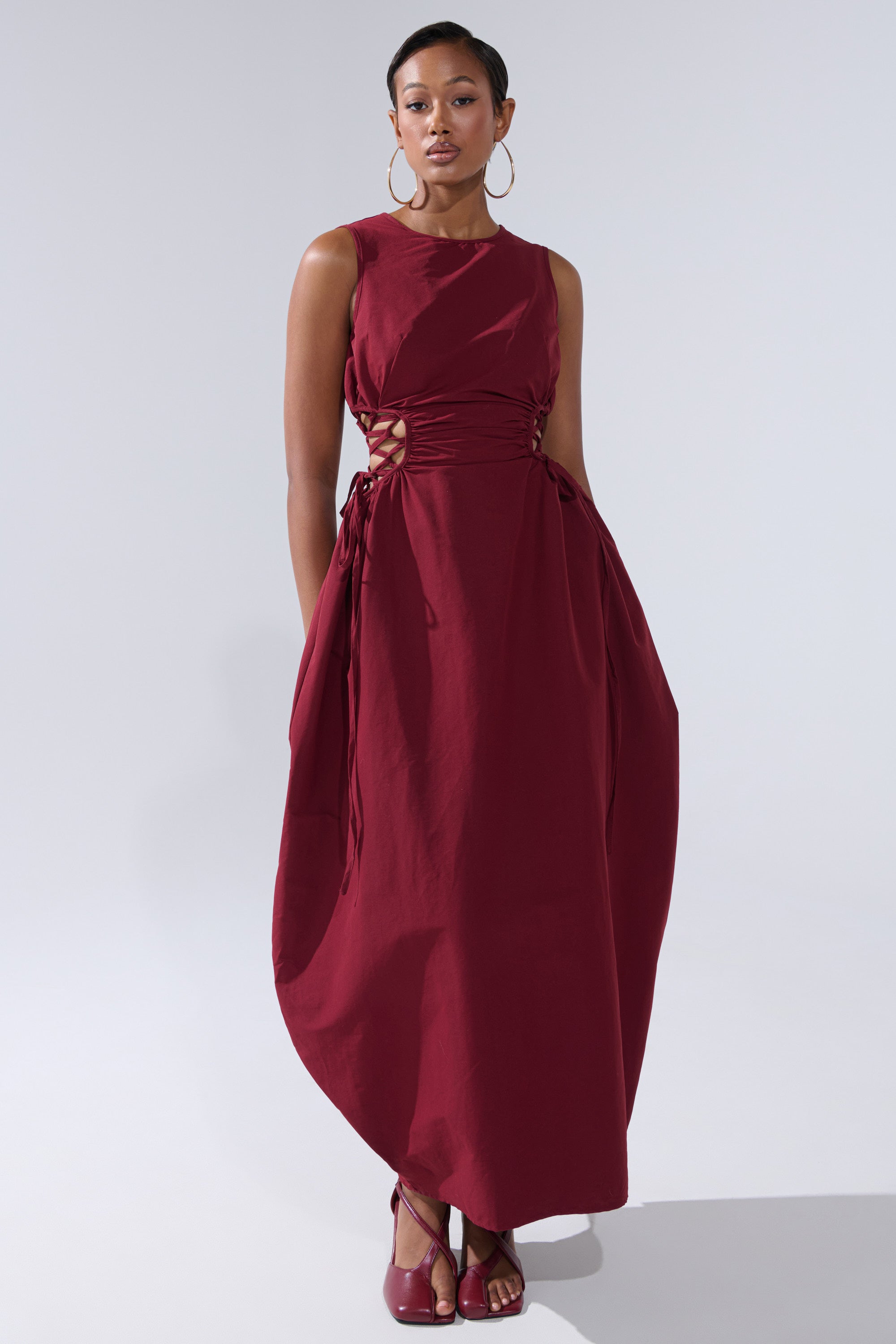 A woman models the ALINA LACE UP DETAIL MAXI DRESS IN BURGUNDY, a sleeveless maxi with side lace-up accents and an asymmetrical hem. She wears hoop earrings, has short hair, and matches with red shoes against a plain light background.