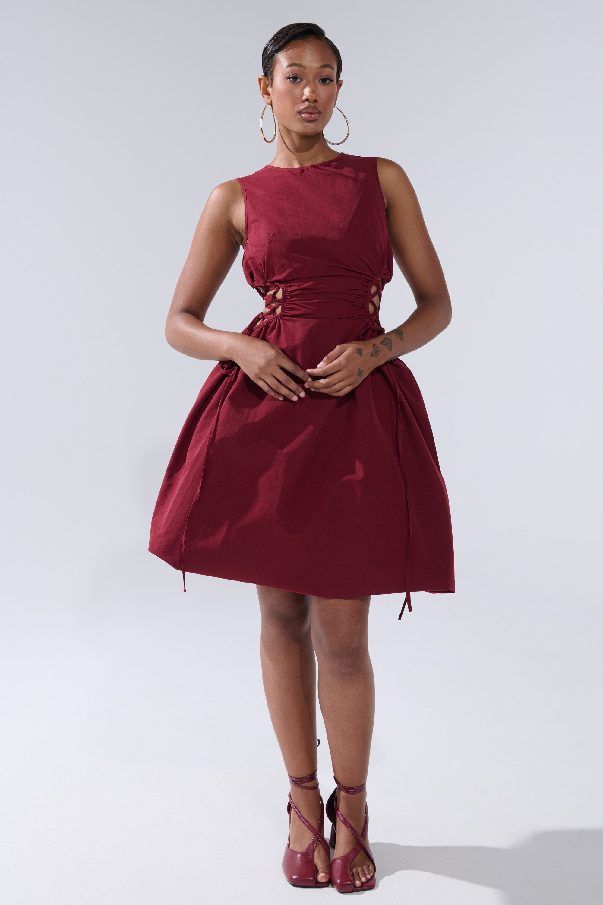 A woman poses against a plain background in the ALINA Lace Up Detail Maxi Dress in Burgundy, styled with matching heels and hoop earrings. Her hair is neatly back, showcasing modern elegance in this standout look.