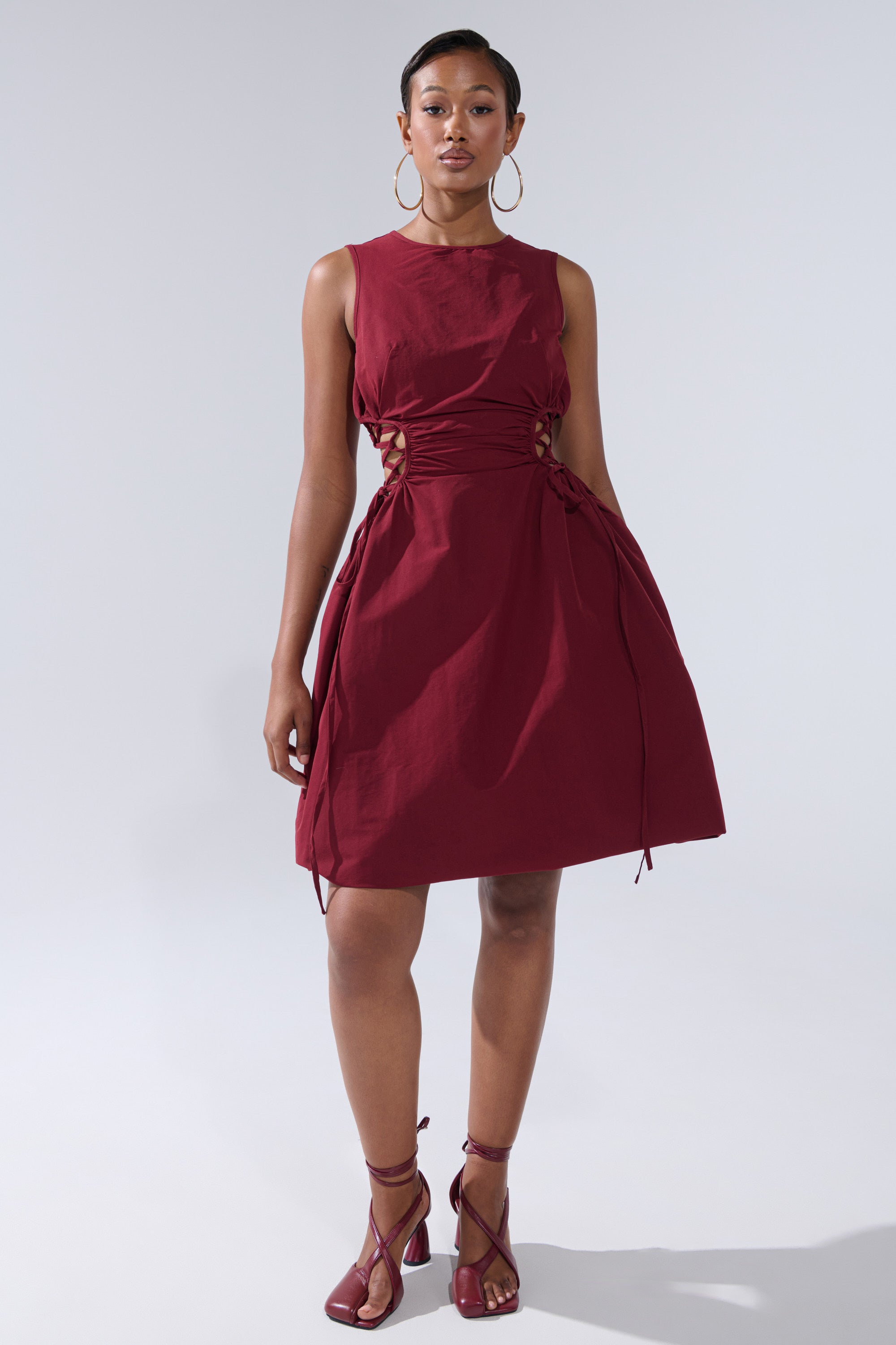 A woman poses against a plain background in the ALINA LACE UP DETAIL MAXI DRESS IN BURGUNDY, styled with matching strappy heels and large hoop earrings.