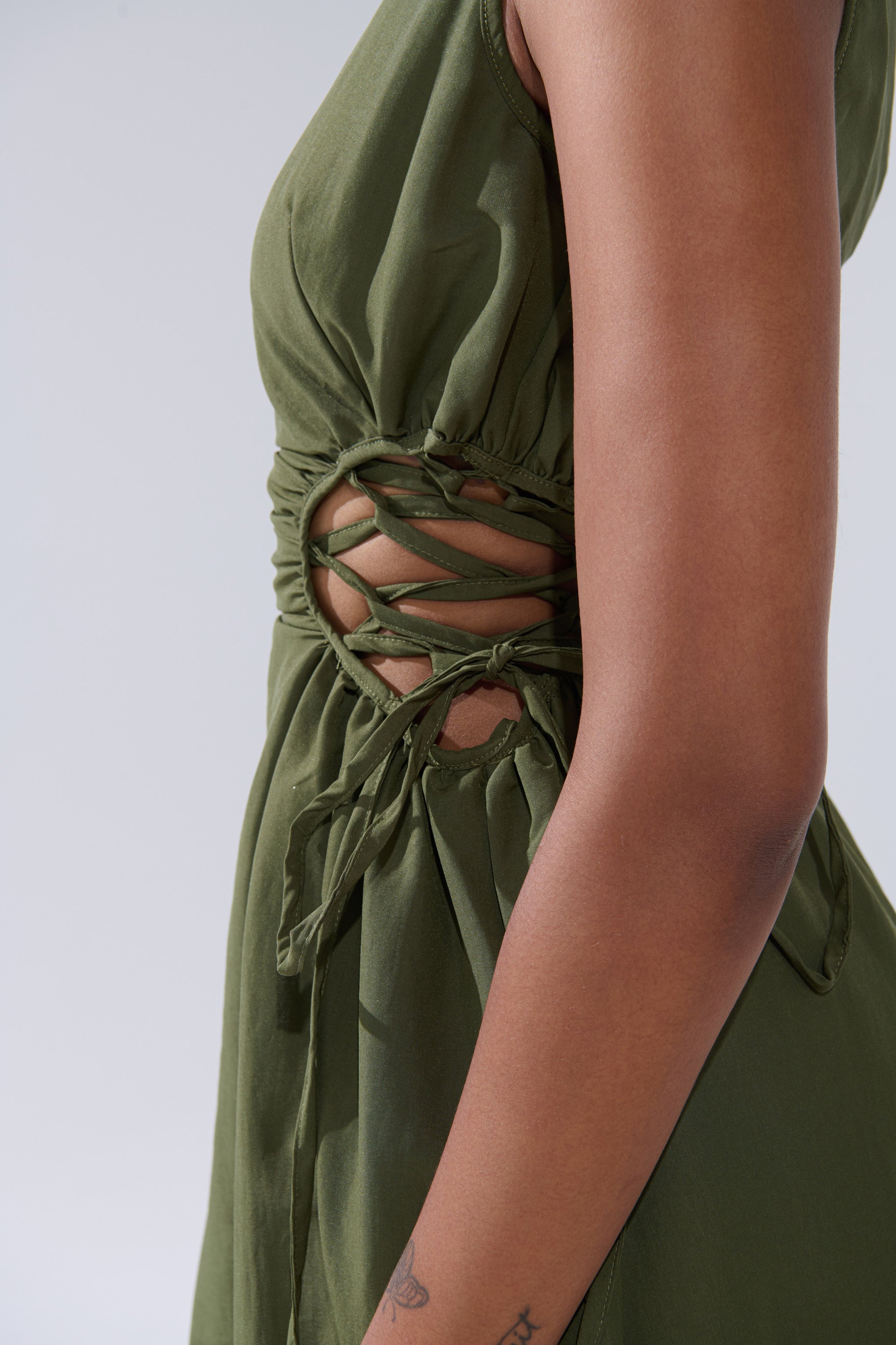 A close-up of a person in the ALINA LACE UP DETAIL MAXI DRESS IN OLIVE, featuring cut-out sides, crisscross lace-up details with bows, and a ruched bodice that reveals their arm and a small forearm tattoo.