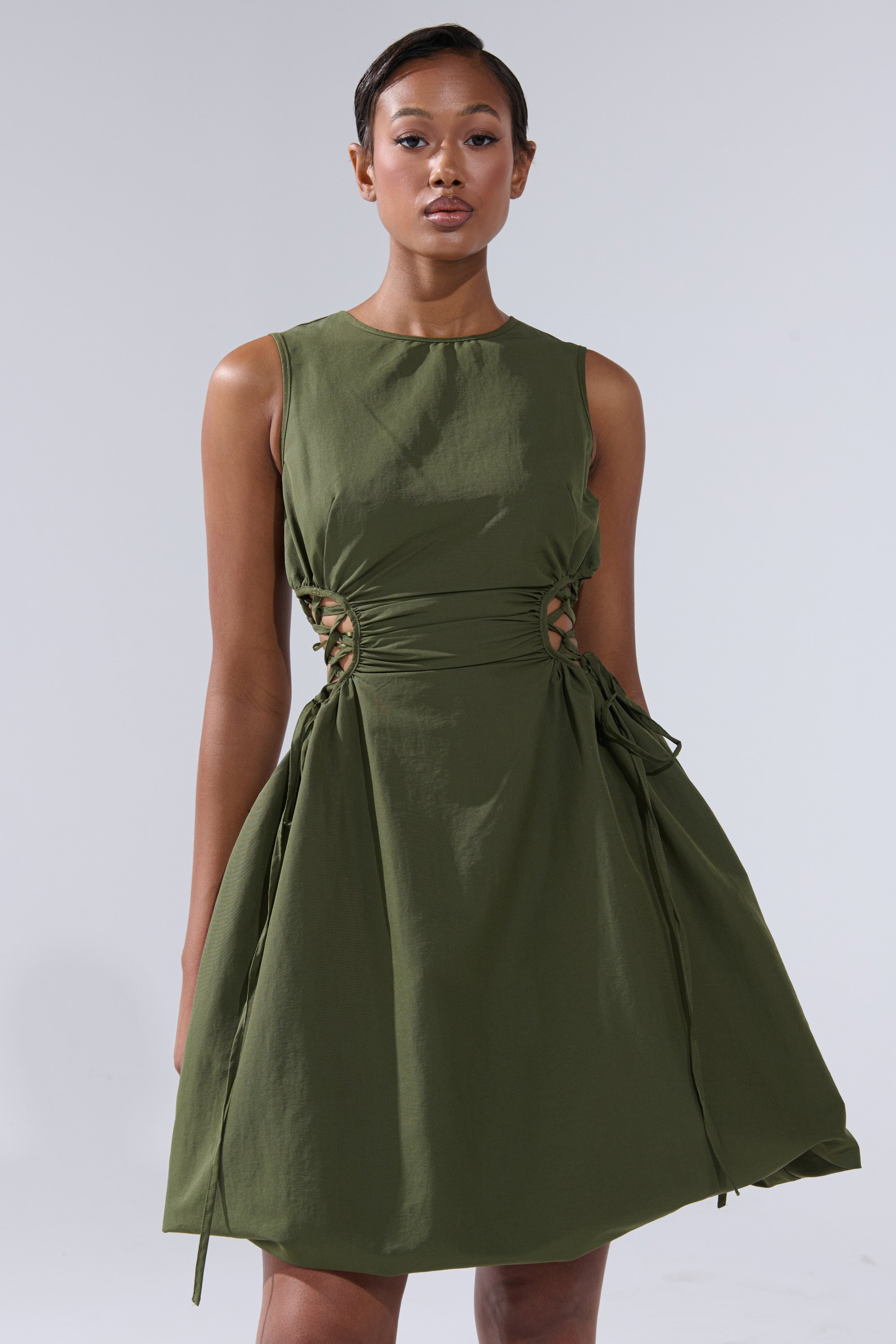 A woman in the ALINA LACE UP DETAIL MAXI DRESS IN OLIVE, a sleeveless olive maxi with side lace-up details, stands confidently against a plain light gray background.
