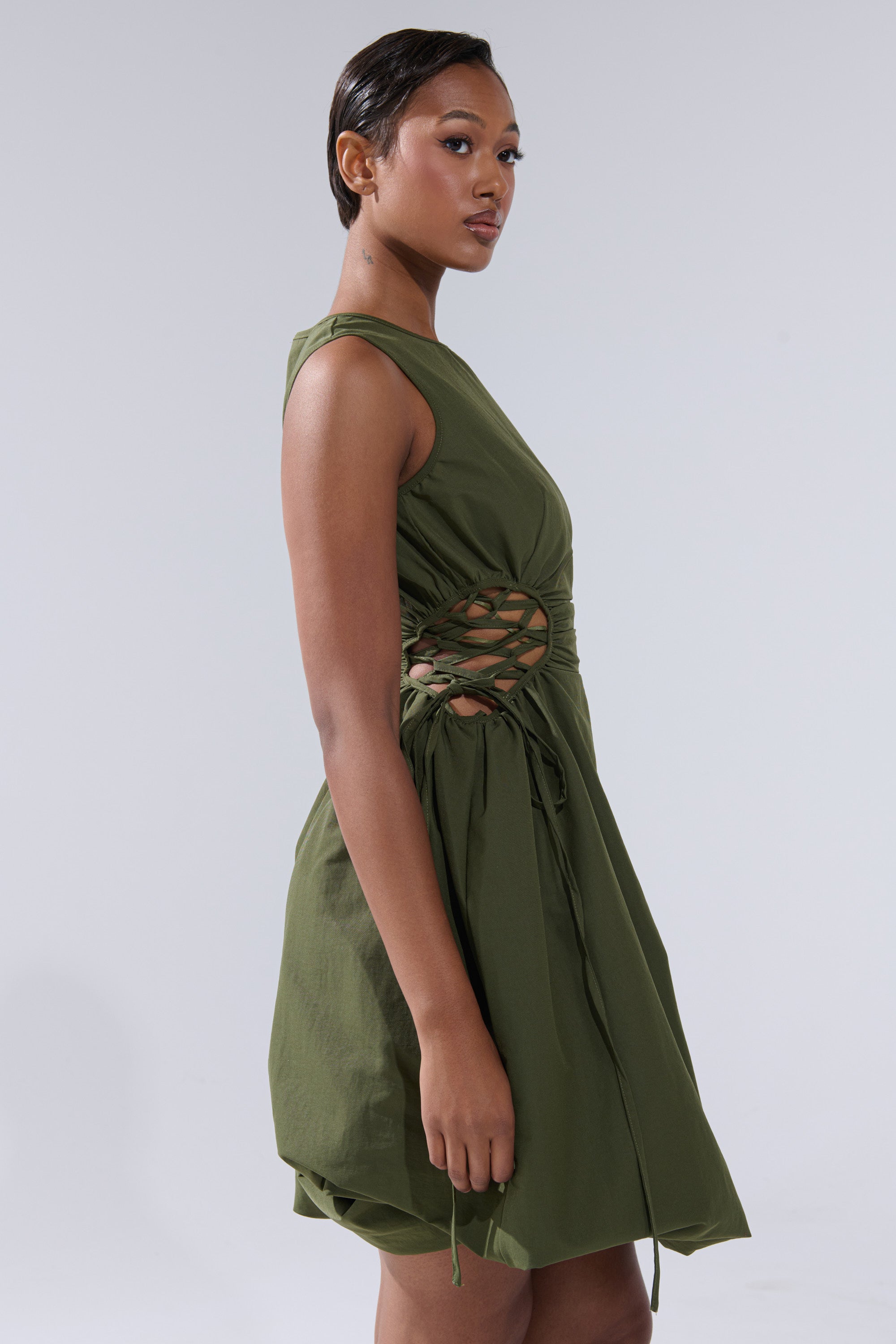 A woman models the ALINA LACE UP DETAIL MAXI DRESS IN OLIVE, featuring a ruched bodice and lattice cutout waist, as she stands confidently against a plain light gray background.