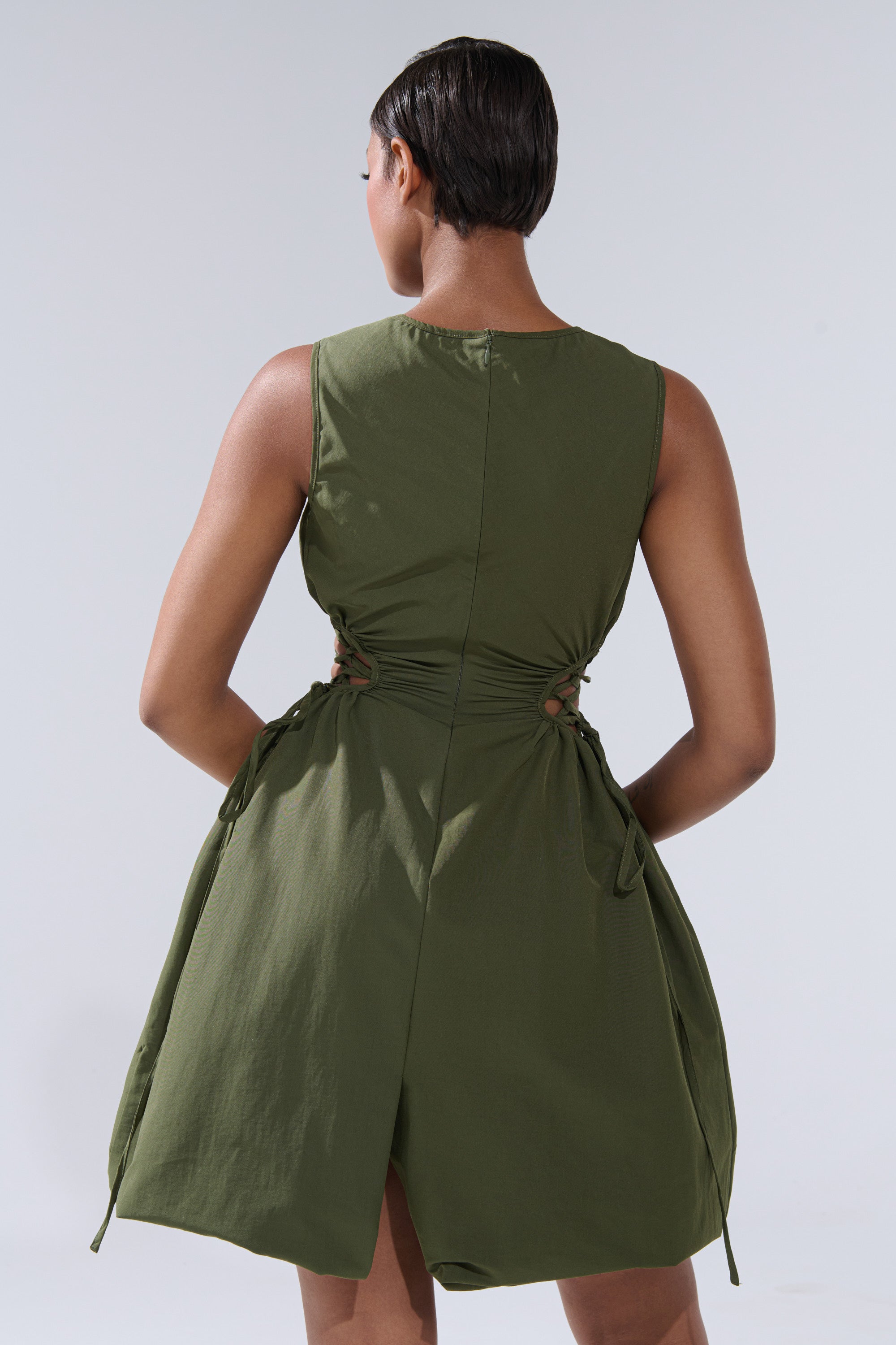 A person with short hair, seen from the back, wears the ALINA LACE UP DETAIL MAXI DRESS IN OLIVE with gathered sides and a slight flare at the hem against a light gray background.
