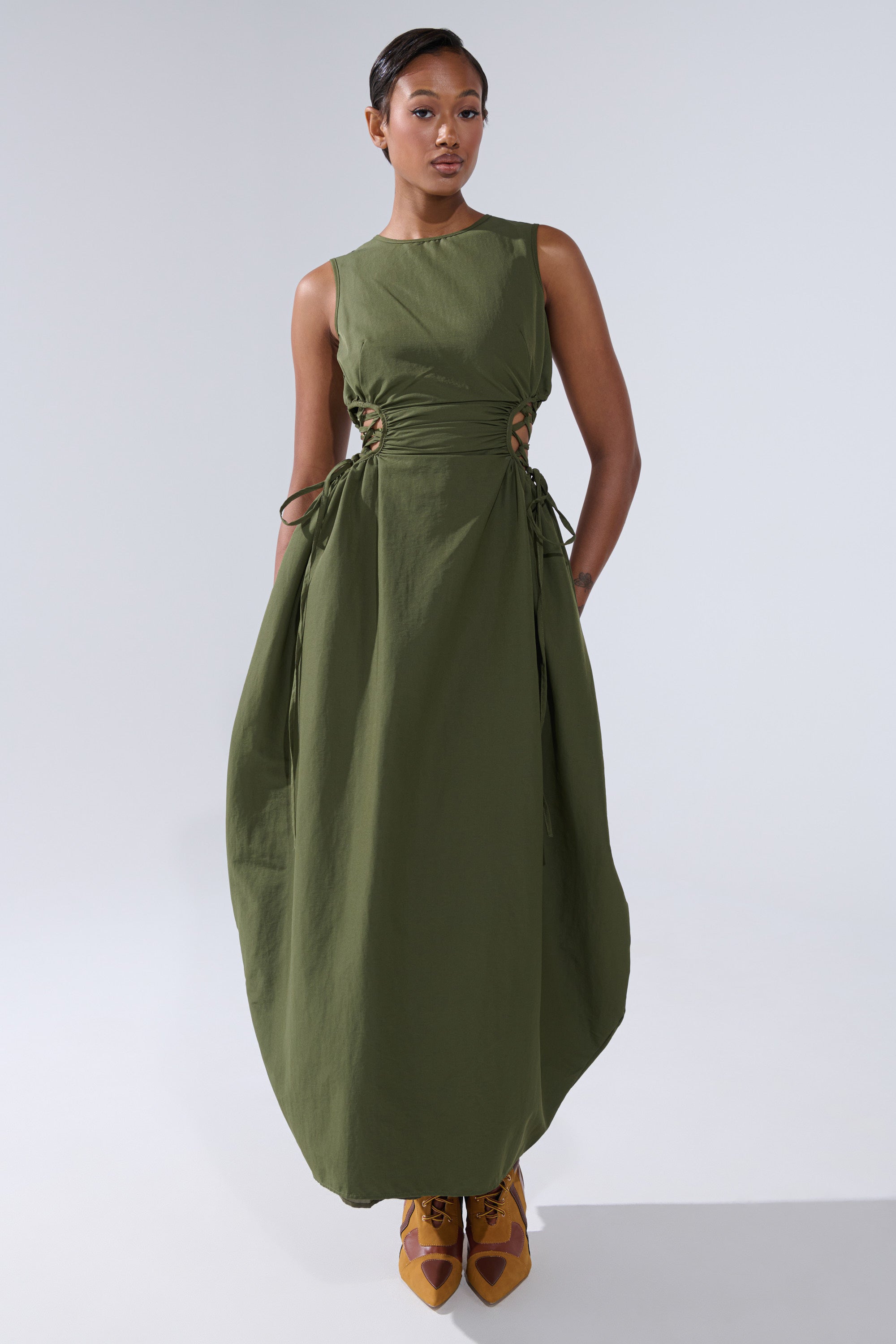 A woman in the ALINA LACE UP DETAIL MAXI DRESS IN OLIVE, featuring side cut-outs and a ruched bodice, pairs it with tan lace-up shoes while standing against a plain light background.