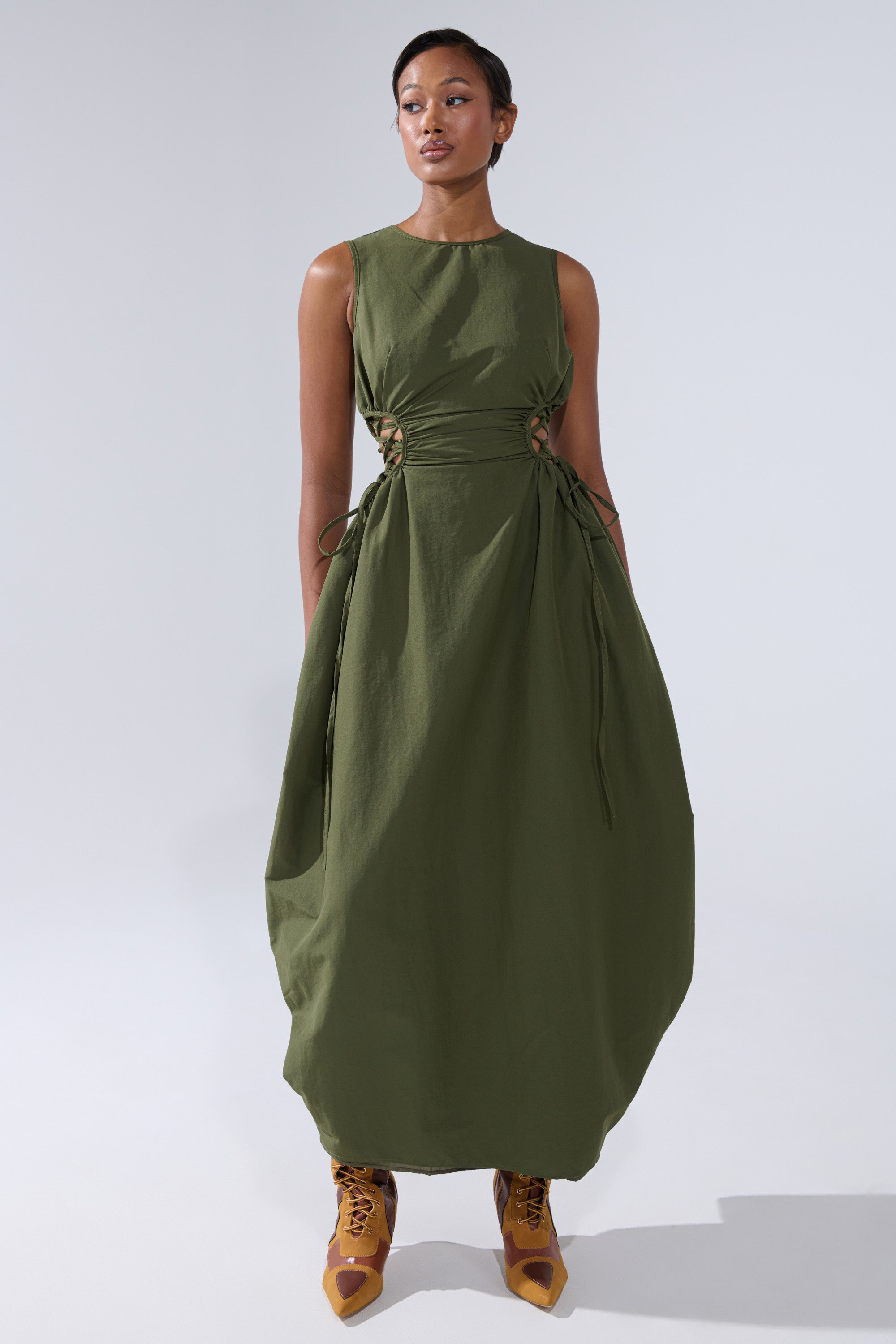 A woman models the ALINA LACE UP DETAIL MAXI DRESS IN OLIVE, featuring a ruched bodice and side ties, paired with mustard yellow lace-up shoes, standing before a plain light gray background.