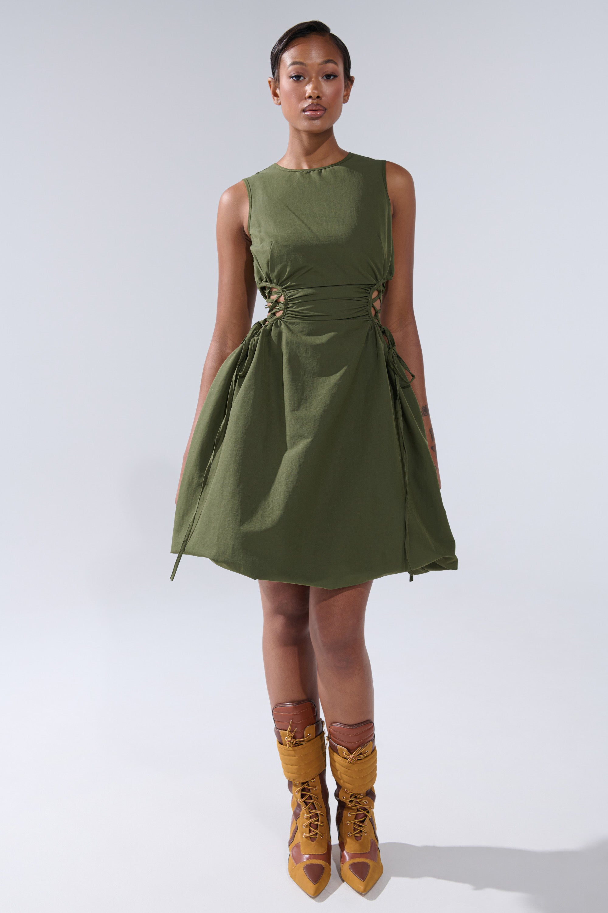 A woman with short hair and a neutral expression faces forward in the ALINA LACE UP DETAIL MAXI DRESS IN OLIVE, paired with mustard yellow lace-up boots, against a plain light background.