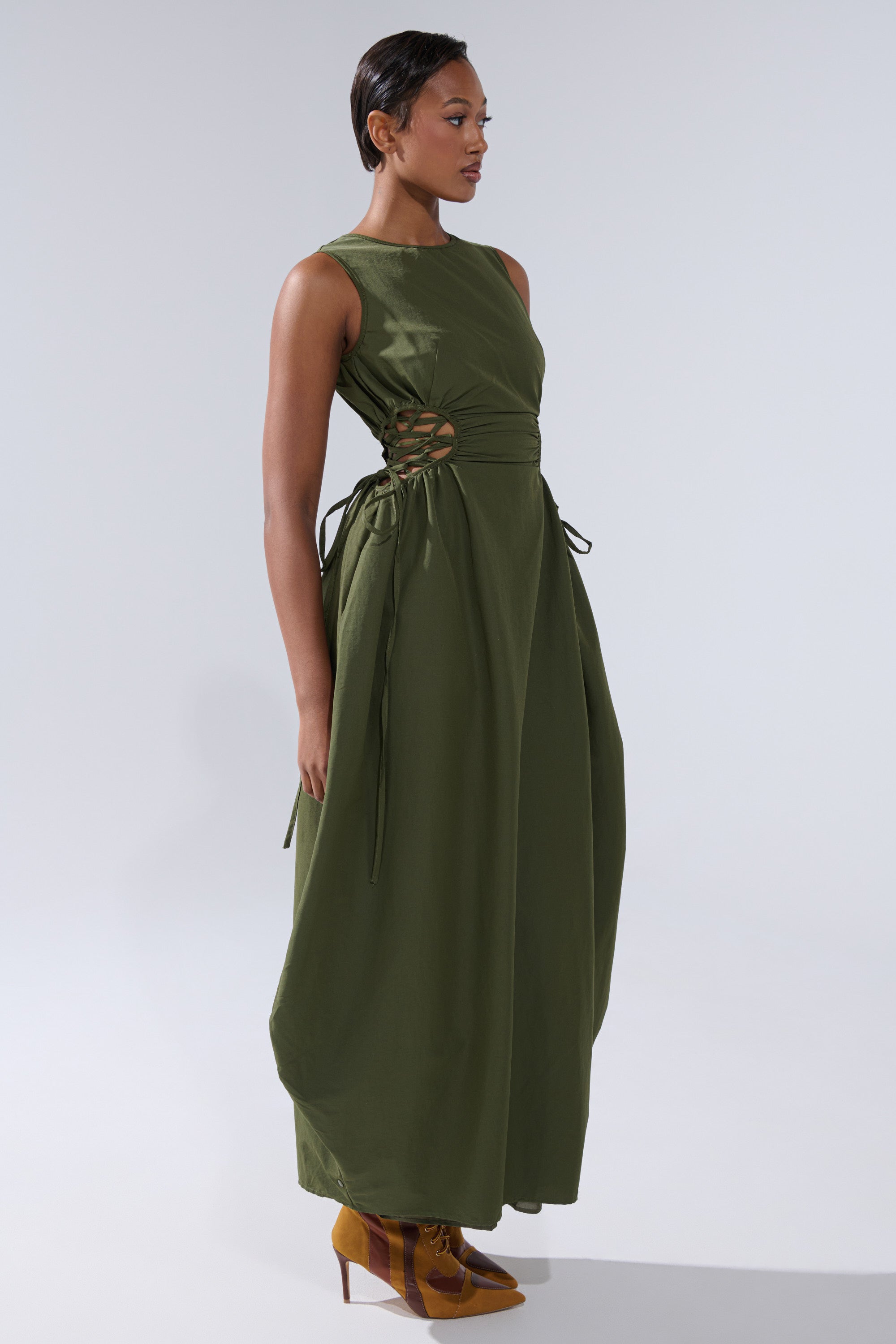 A woman wears the ALINA LACE UP DETAIL MAXI DRESS IN OLIVE—an olive maxi dress featuring a ruched bodice and side lace-up details—paired with brown lace-up shoes, standing against a plain, light background.