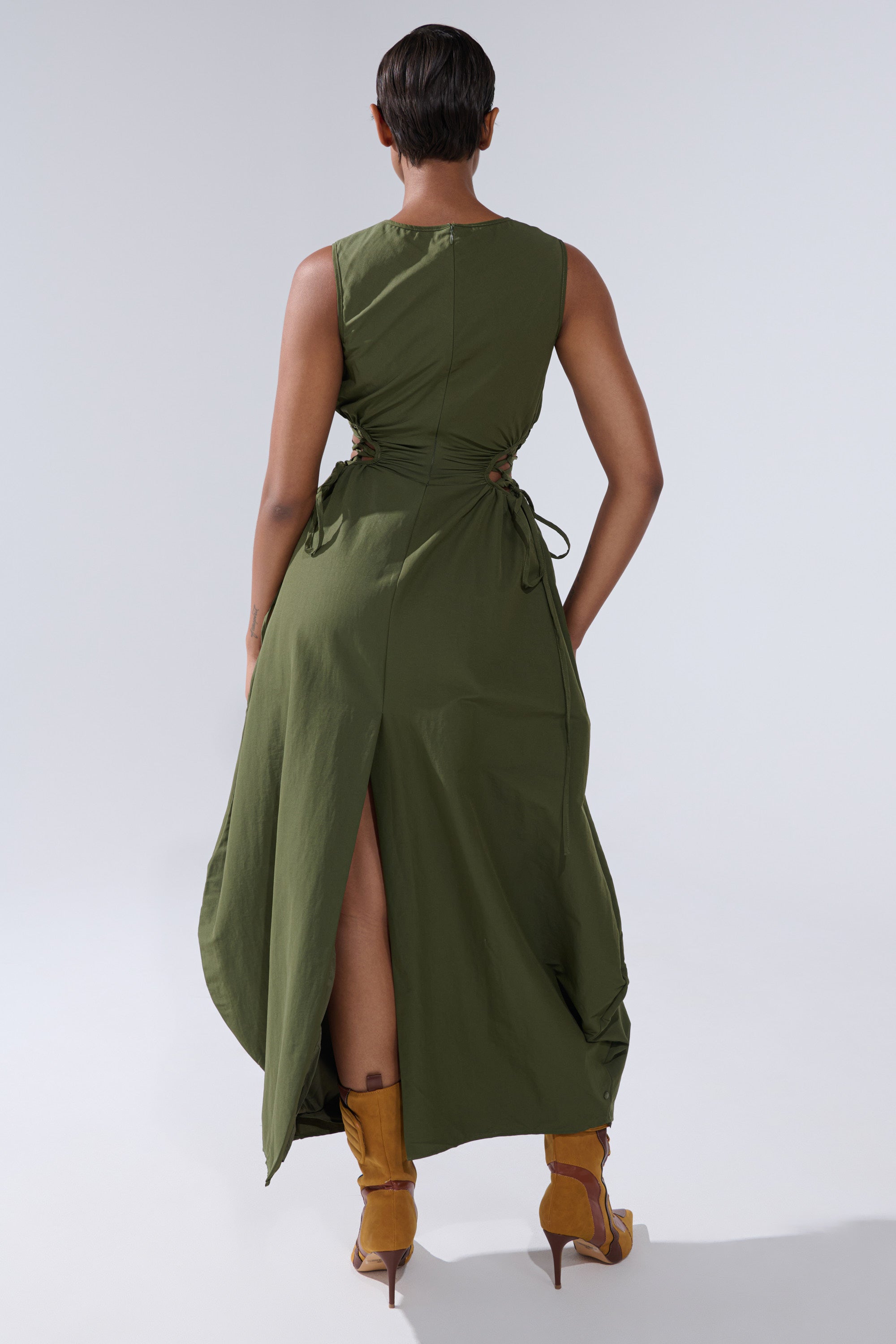 Wearing the ALINA LACE UP DETAIL MAXI DRESS IN OLIVE, featuring side ties and a back slit, paired with tan high-heeled boots, a person faces away from the camera against a plain light background.