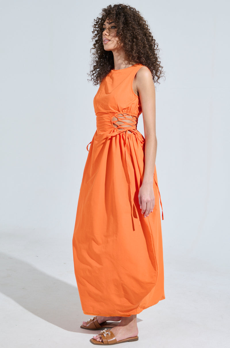 A woman with curly hair stands in profile, wearing the ALINA Lace Up Detail Maxi Dress in orange, featuring lace-up side tie details and a flowing skirt. She pairs it with brown sandals against a plain, light-colored backdrop.