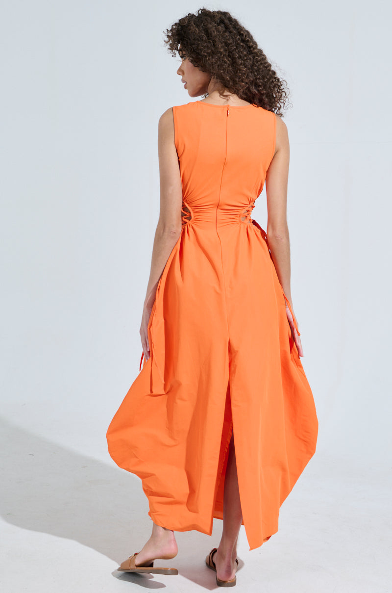A person with curly hair stands wearing the ALINA Lace Up Detail Maxi Dress in orange, a sleeveless design made from a soft cotton blend with side ties and a back slit. They're facing away from the camera against a light gray background.