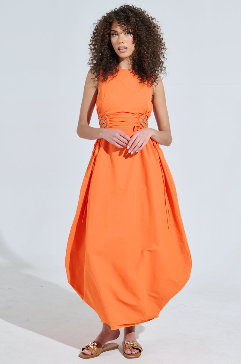 A woman with curly hair stands against a light background, wearing the ALINA LACE UP DETAIL MAXI DRESS IN ORANGE, featuring a convertible silhouette and balloon hem. She pairs it with brown slip-on sandals, exuding a relaxed posture.