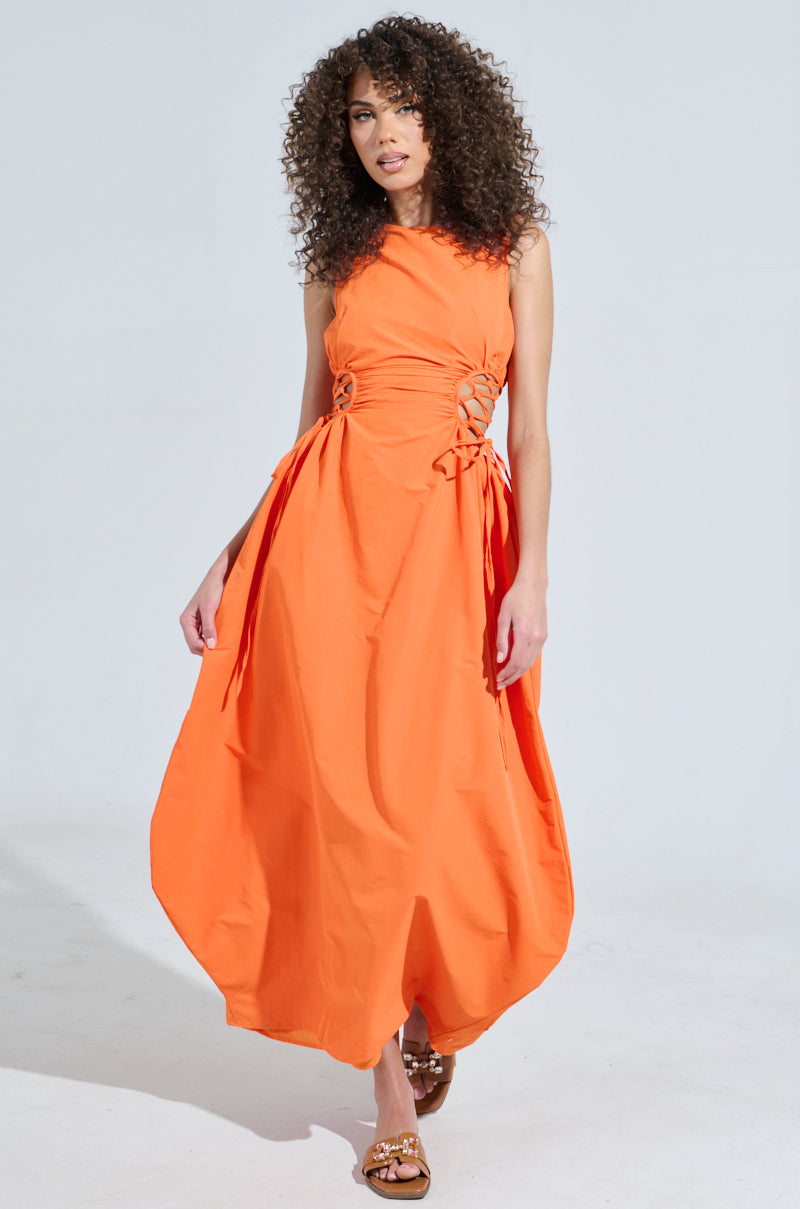 A person with curly hair is wearing the ALINA LACE UP DETAIL MAXI DRESS IN ORANGE and brown sandals, standing against a plain background.