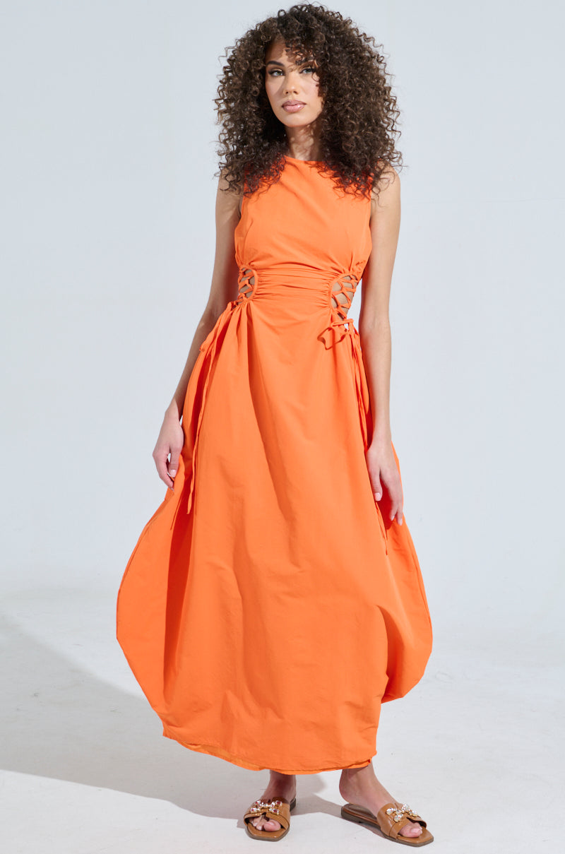 A person with curly hair wears the ALINA LACE UP DETAIL MAXI DRESS IN ORANGE, standing against a light background, paired with brown sandals.