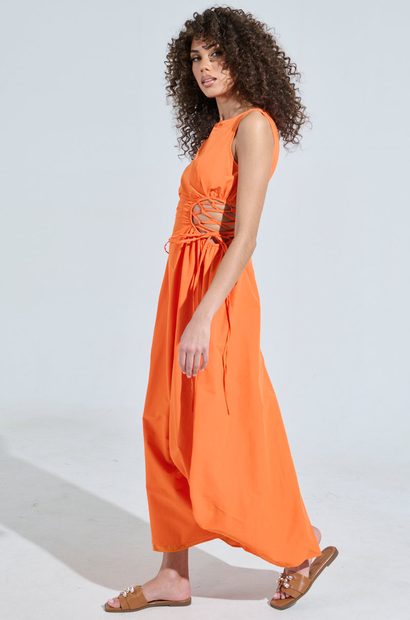 With curly hair, a person wears the ALINA LACE UP DETAIL MAXI DRESS IN ORANGE, sleeveless and elegantly designed with lace-up sides, paired with brown sandals. They pose gracefully against a plain light-colored background.
