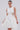 A woman wears the ALINA LACE UP DETAIL MAXI DRESS IN WHITE, featuring a sleeveless design, fitted bodice, and A-line skirt. Her hair is pulled back, complemented by a silver necklace against a plain, light background.