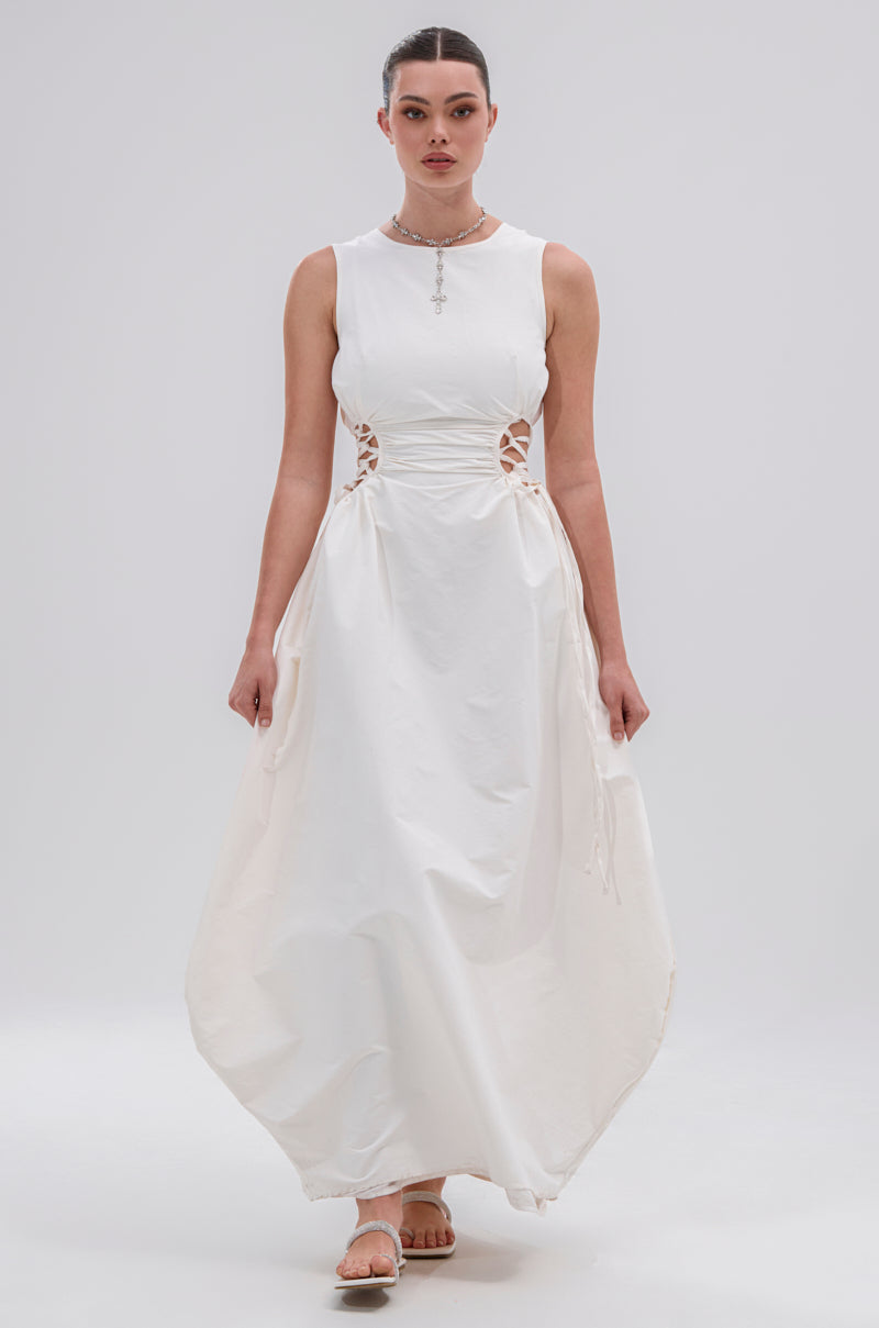 A woman stands facing forward against a plain light background, wearing the ALINA Lace Up Detail Maxi Dress in White with side lace-up details and a flared hem. Her hair is tied back, and she accessorizes with sandals and a necklace.