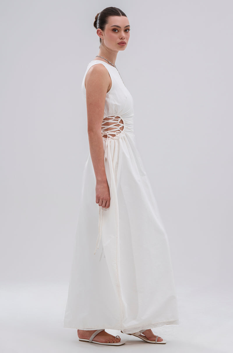 A woman stands sideways wearing the ALINA LACE UP DETAIL MAXI DRESS IN WHITE, a sleeveless, floor-length dress with side detailing. Her hair is styled in a neat bun, and she pairs the look with white sandals against a plain light backdrop.