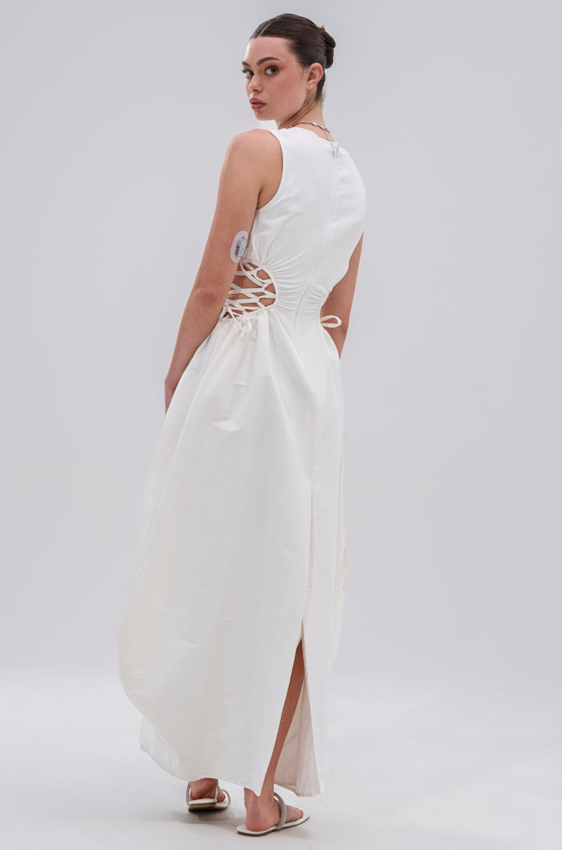 A woman with her hair in a bun faces away, wearing the ALINA LACE UP DETAIL MAXI DRESS IN WHITE with a back slit and white sandals. The background is plain and light-colored.