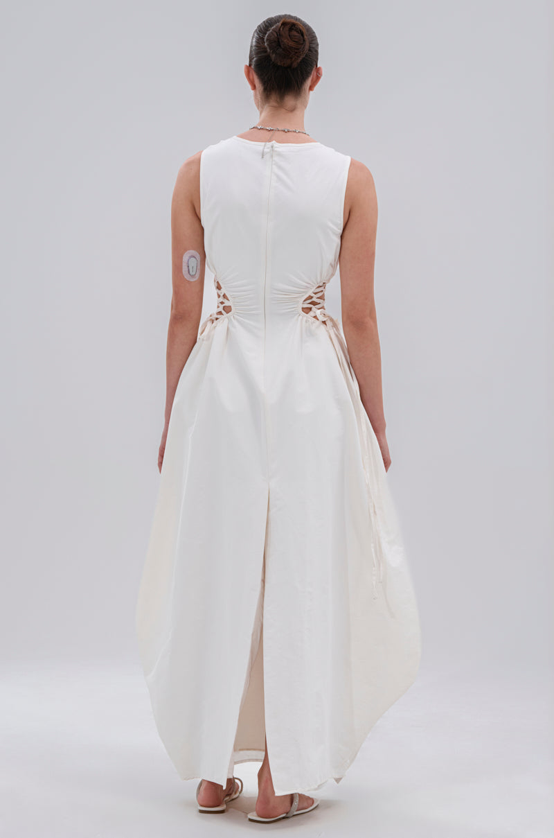 A person faces away, wearing the ALINA LACE UP DETAIL MAXI DRESS IN WHITE with lace-up sides and a back slit. Their hair is in a bun, they wear sandals, and a circular medical patch is on their upper left arm.
