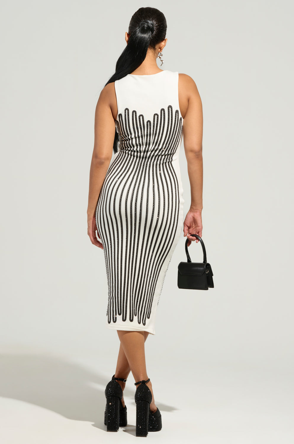 The individual is seen from the back wearing the ITS GIVING BODY RHINESTONE PRINTED MIDI DRESS, which features a sleeveless design with vertical black stripe patterns that enhance their figure. They accessorize with a small black handbag and are elevated by high platform heels, all set against a plain gray background.