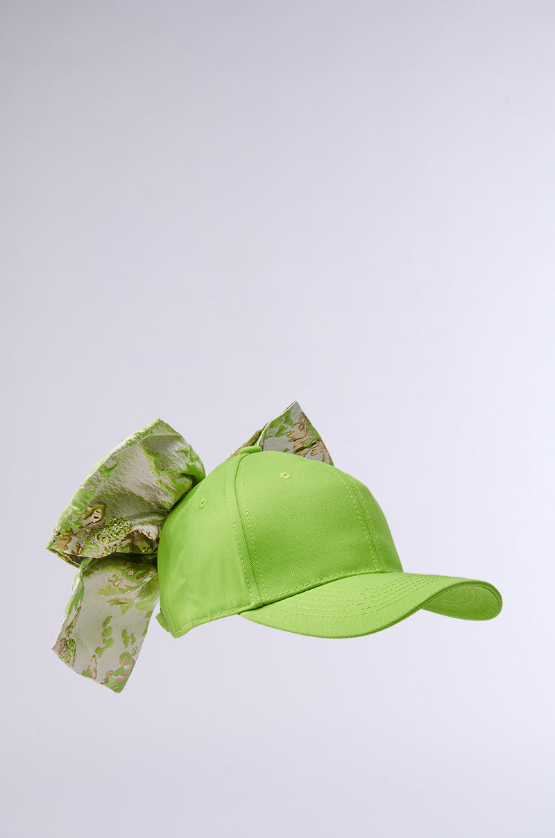 The DIVINE DREAMS BROCADE BOW HAT is a lime green cap with an oversized, decorative floral-patterned ribbon featuring soft green and beige tones at the back, adding elegance to its casual design.