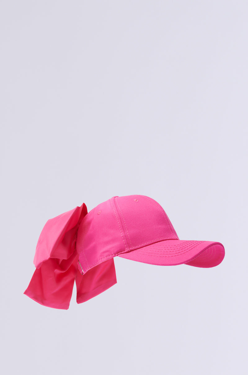 The VALENTINA BOW HAT is a vibrant pink baseball cap with an adjustable buckle and a large matching fabric bow attached to the back, set on a light gray background.