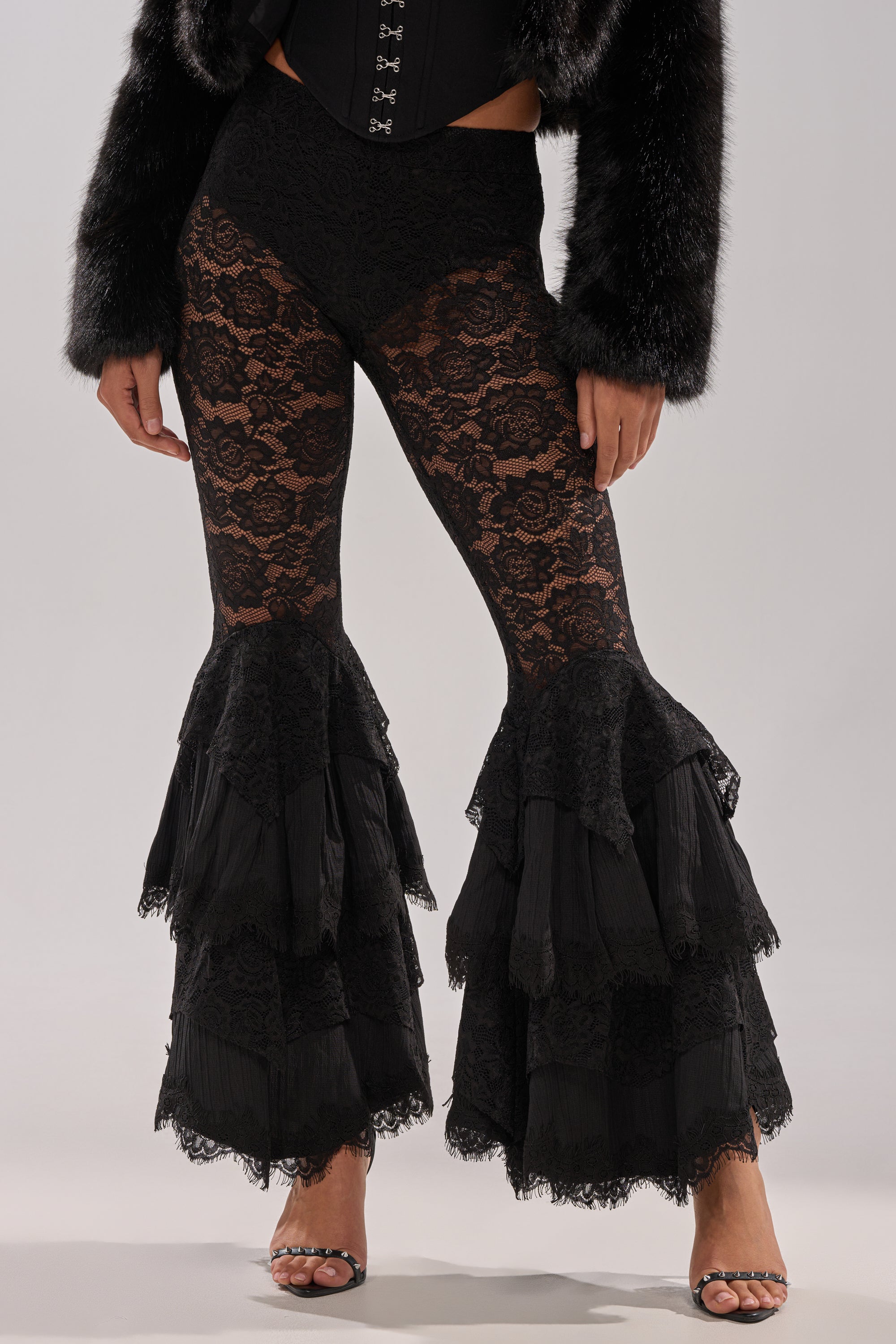 A person models the COACHELLA LACE RUFFLE PANTS with flared, tiered bottoms, paired with a black faux fur jacket and heels—a striking festival look that stands out against a plain background.
