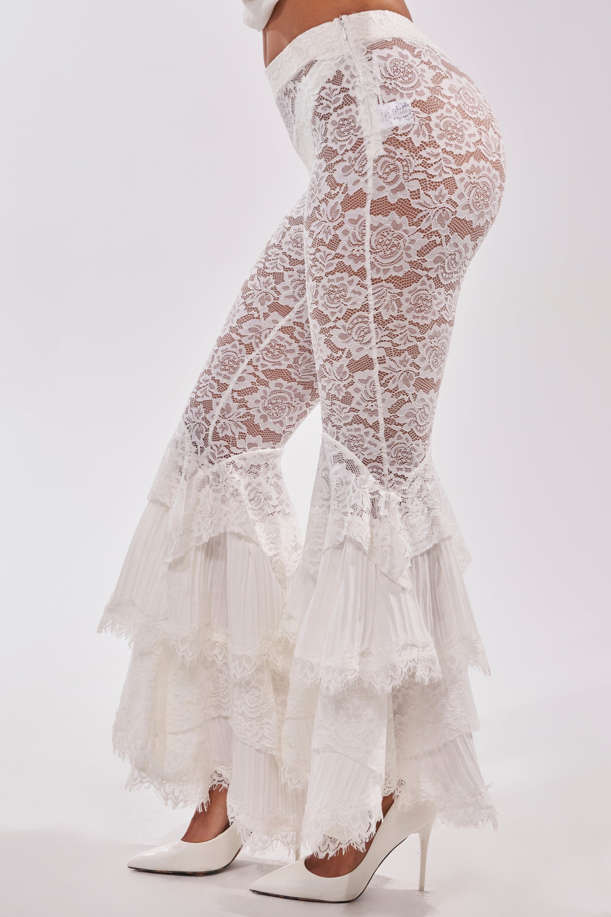 A person models COACHELLA LACE RUFFLE PANTS with high waist, tiered flares, and white heels. The cropped image focuses on the lower torso and legs against a white background, emphasizing their festival-ready boho style. 3