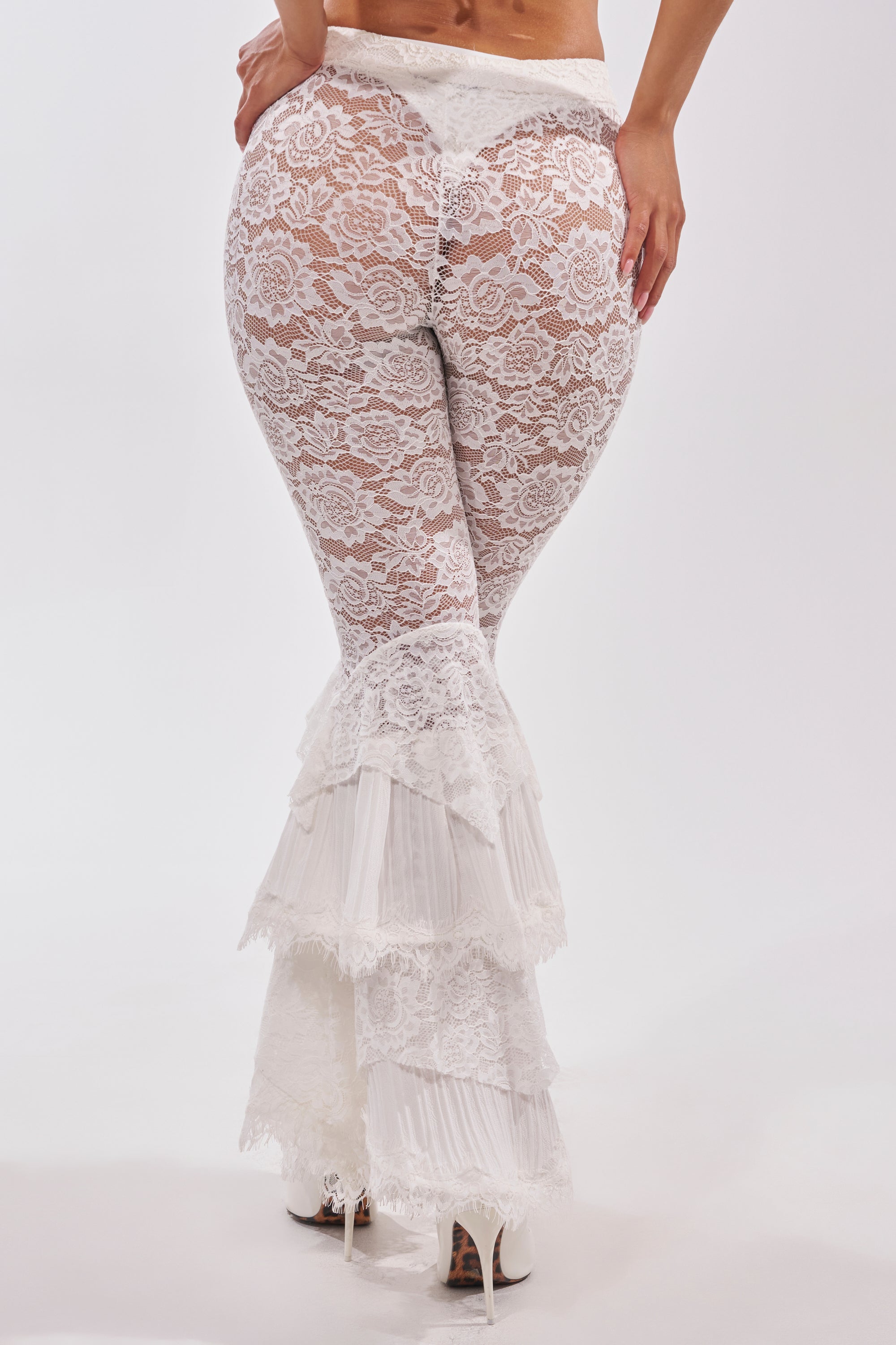 Wearing the COACHELLA LACE RUFFLE PANTS—white boho ruffle pants with sheer lace bell-bottoms and ruffled tiers—a person in high heels stands with their back to the camera against a plain white background. 4