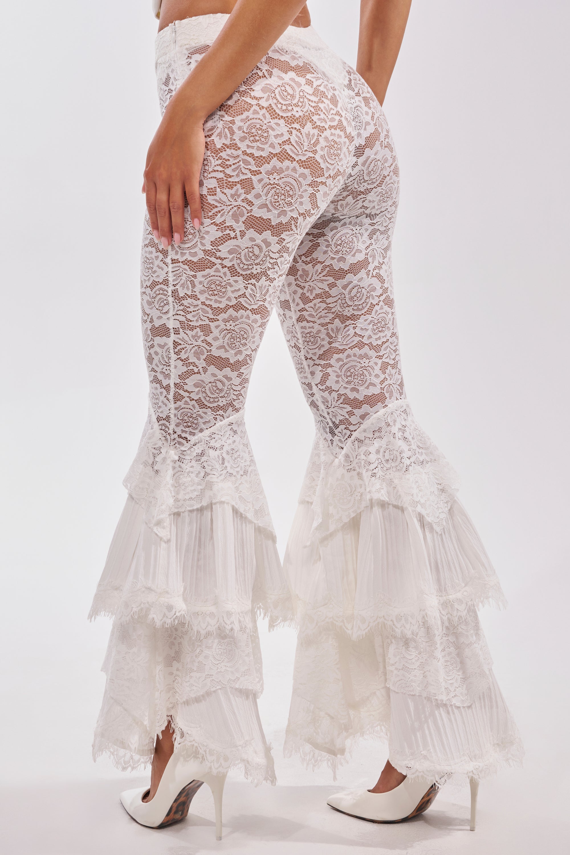 A person wearing COACHELLA LACE RUFFLE PANTS with layered lace ruffles at the bottom and white heels stands against a plain white background. 5