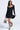 A woman models the IN THE MOOD BANDAGE MINI DRESS, a fitted, asymmetrical long-sleeve mini dress. She holds the hem, accessorized with a leopard print handbag and black strappy heels, posing against a plain white background.