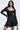 A woman stands confidently against a plain background, wearing the IN THE MOOD BANDAGE MINI DRESS—a fitted, off-shoulder black mini with long sleeves, a sweetheart neckline, and a flared skirt—looking elegantly to the side.