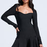 A woman stands confidently against a plain background, wearing the IN THE MOOD BANDAGE MINI DRESS—a fitted, off-shoulder black mini with long sleeves, a sweetheart neckline, and a flared skirt—looking elegantly to the side.
