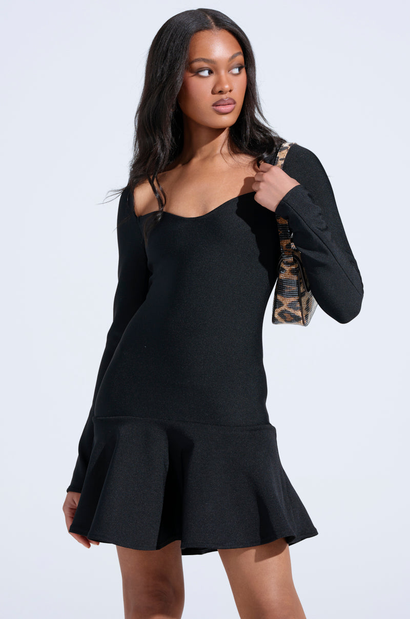 A woman with long dark hair wears the IN THE MOOD BANDAGE MINI DRESS—a black, fitted, long-sleeve mini dress with a sweetheart neckline and flared skirt—while holding a patterned shoulder bag against a plain light background.