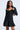 A woman with long dark hair wears the IN THE MOOD BANDAGE MINI DRESS—a black, fitted, long-sleeve mini dress with a sweetheart neckline and flared skirt—while holding a patterned shoulder bag against a plain light background.