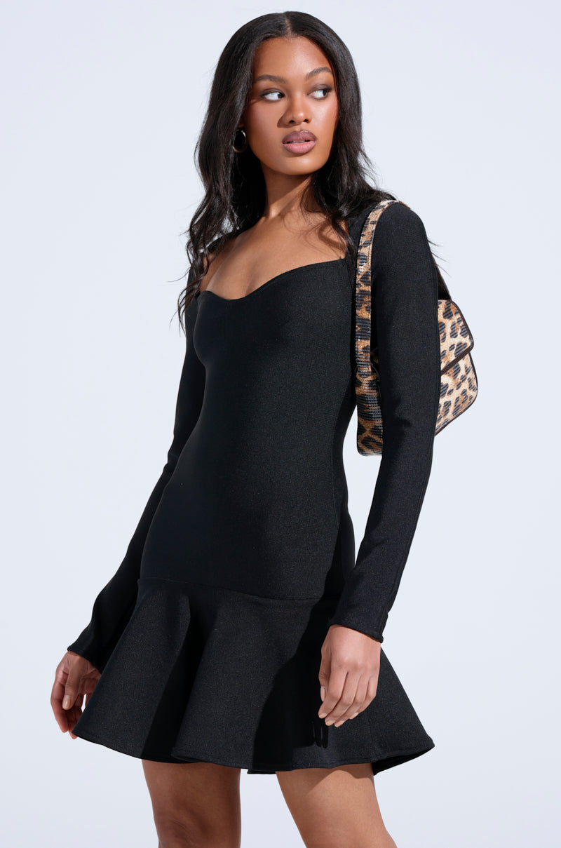 A woman stands against a plain light background, wearing the fitted black AKIRA Label "IN THE MOOD BANDAGE MINI DRESS" with long sleeves and a flared hem, paired with a leopard-print shoulder bag.