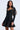 A woman stands against a plain light background, wearing the fitted black AKIRA Label 