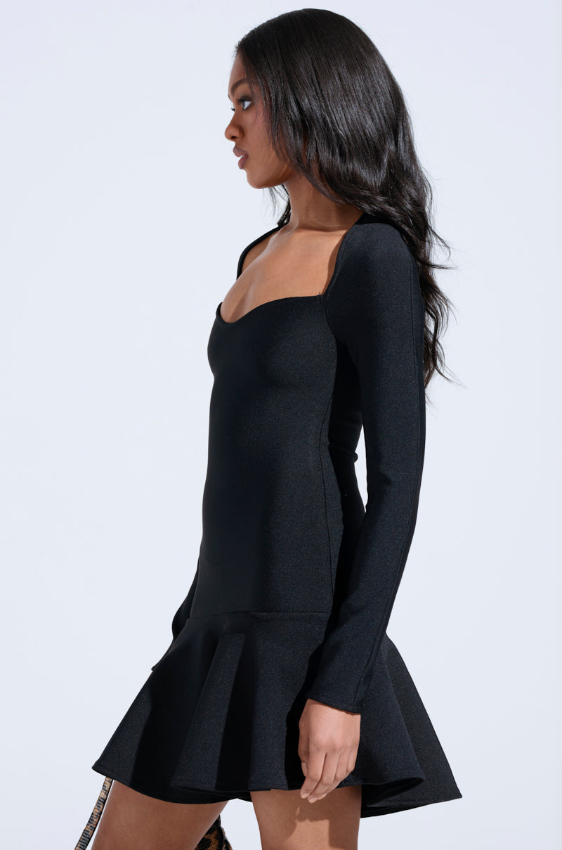 A woman with long, wavy dark hair wears the IN THE MOOD BANDAGE MINI DRESS—fitted, with a flared skirt and long sleeves—as she stands sideways against a plain light background, gazing ahead.