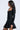 A woman with long, wavy dark hair wears the IN THE MOOD BANDAGE MINI DRESS—fitted, with a flared skirt and long sleeves—as she stands sideways against a plain light background, gazing ahead.