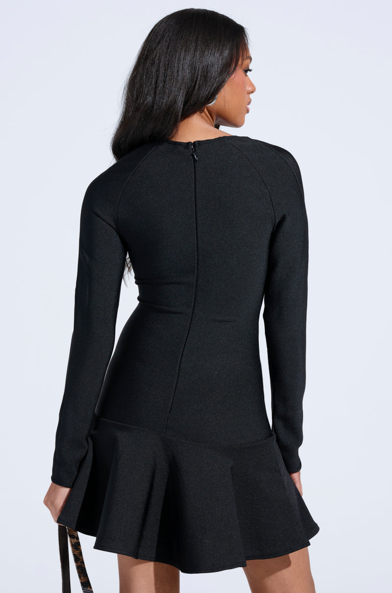 A woman with long dark hair is seen from the back wearing the IN THE MOOD BANDAGE MINI DRESS, a fitted mini with long sleeves and a flared skirt, holding a small bag in her left hand against a plain white background.